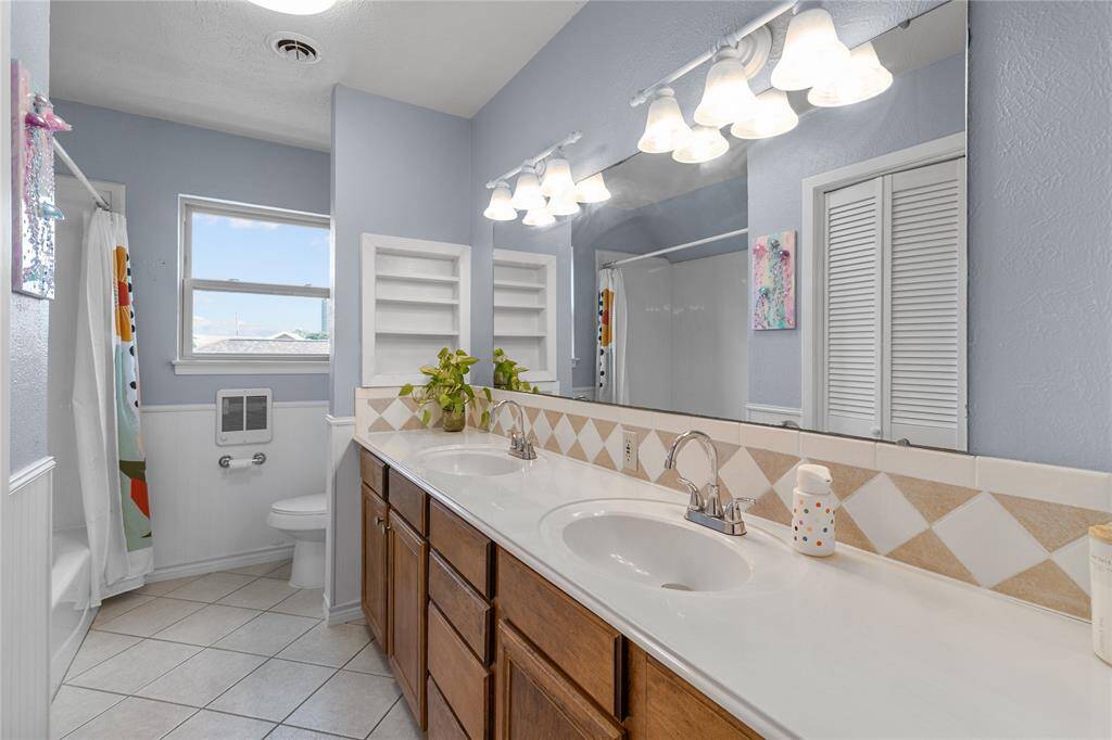 This bathroom features a double-sink vanity with ample storage, a large mirror, and overhead lighting. It has a tiled floor and a shower-tub combo. The space is brightened by a window and includes built-in shelves for storage.