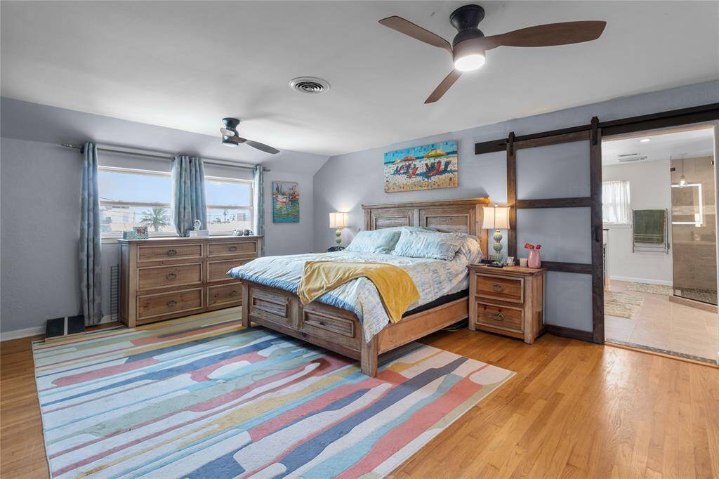 Spacious bedroom with hardwood floors.  It has large windows, very spacious and bright and new ceiling fans. The room includes a sliding barn door leading to an en-suite bathroom, creating a modern and inviting atmosphere.