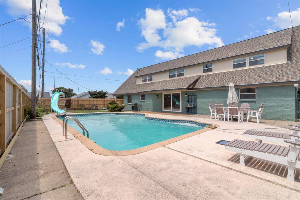 This spacious backyard features a large in-ground pool with a slide, surrounded by a concrete deck. There's outdoor seating and a dining area, perfect for entertaining. The home has a unique, sloped roof design and multiple windows for natural light.