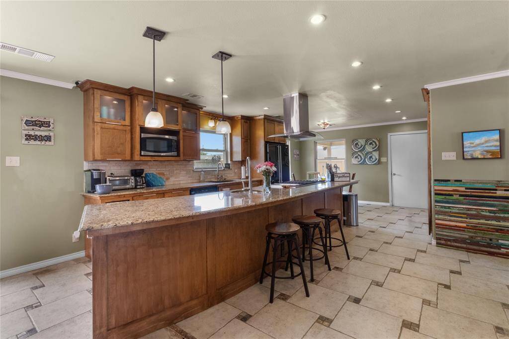 This spacious kitchen features a large granite island with bar seating, modern pendant lighting, and ample wooden cabinetry. It includes stainless steel appliances, a cozy breakfast nook, and a unique tile floor design, creating a warm and inviting atmosphere.