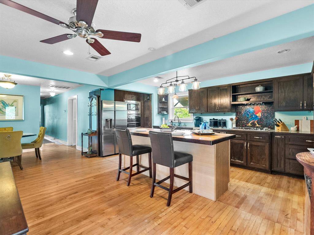 This open-concept kitchen and living area features light blue walls, hardwood floors, and a central island with pendant lighting. It includes modern stainless steel appliances, a cozy seating area, and ample natural light from large windows, creating an inviting and functional space.