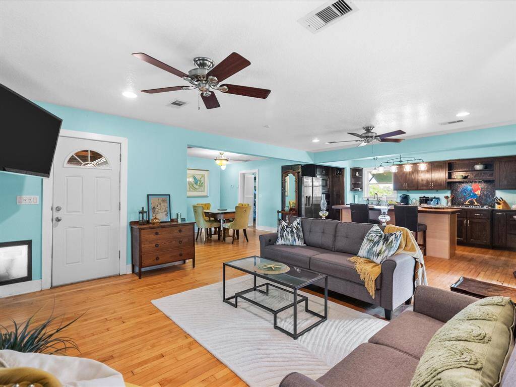 This spacious, open-concept living area features a bright, airy design with light blue walls and hardwood floors. The room includes ceiling fans, modern furnishings, and a seamless flow into the dining and kitchen areas, which boast dark cabinetry and ample natural light.