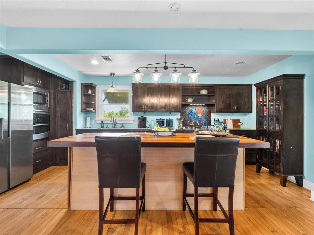 This kitchen features a spacious island with seating, modern stainless steel appliances, and dark wood cabinetry. It has a light blue wall color, pendant lighting, and ample natural light from a window above the sink, creating a warm and inviting space.