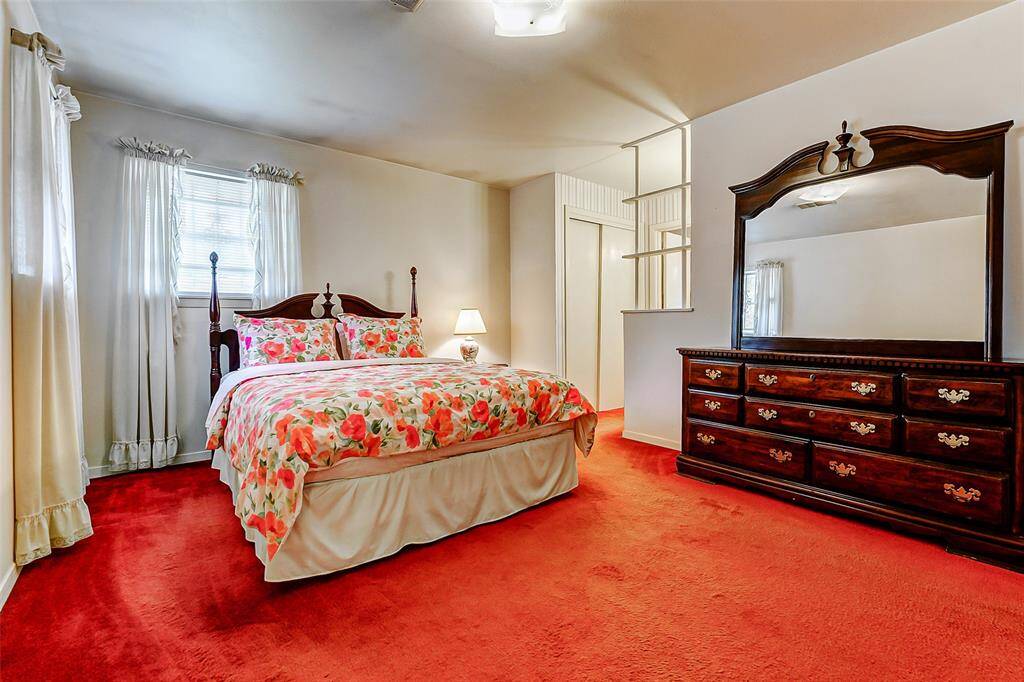 Charming primary bedroom with vintage character, highlighted by bright natural light, fresh white walls, and plush red carpet. An oversized mirror and classic furniture styling add a timeless, elegant touch.