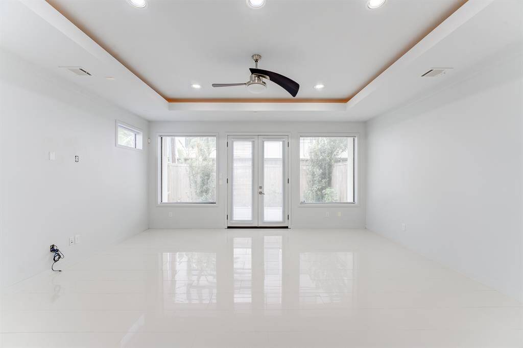 Bright, spacious room with sleek white flooring, modern recessed lighting, and a central ceiling fan. Large windows and glass doors offer ample natural light, leading to an outdoor view. Ideal for a contemporary living area.