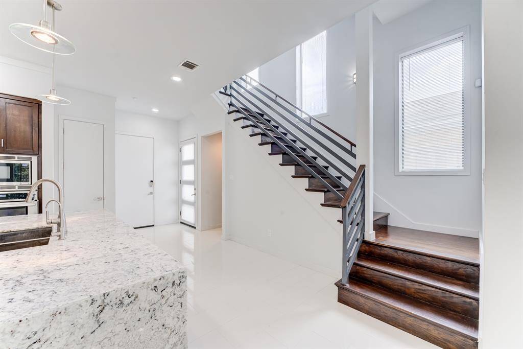 This photo showcases a modern, open-concept kitchen and entryway. It features high ceilings and large windows. A stylish staircase with dark wood steps and metal railing leads to the upper level, complemented by elegant light fixtures.