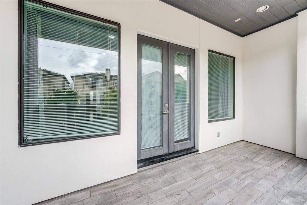 This photo shows a covered patio area with light-colored tile flooring, large windows with blinds, and double glass doors, providing a bright and inviting entryway.