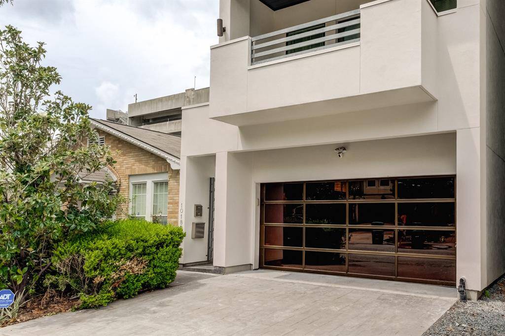Modern home exterior featuring a sleek, glass-paneled garage door and clean lines. The driveway is paved, and there's a small landscaped area with greenery, providing a contemporary and inviting entrance.
