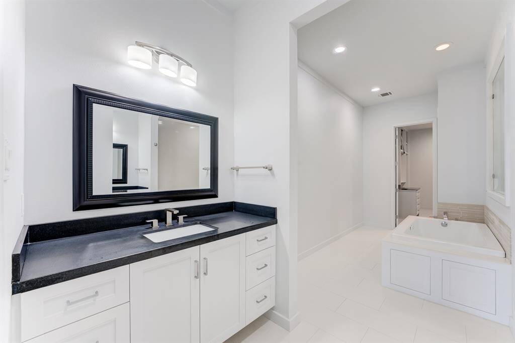 This bathroom features a modern design with a sleek white vanity, black countertop, and large framed mirror. It includes a spacious bathtub and ample lighting, creating a bright and inviting space.