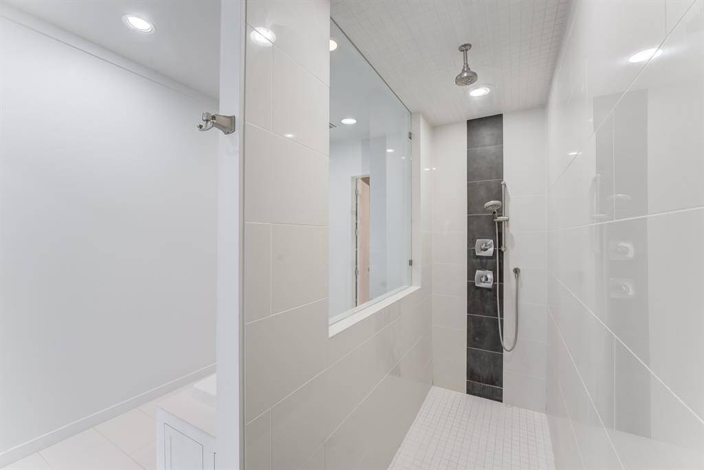 Modern shower with sleek white tiles, a rainfall showerhead, and a glass partition. The space is bright and stylish, featuring a contrasting vertical dark tile strip and recessed lighting.