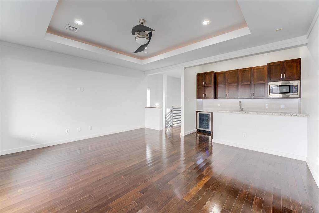 This spacious room features elegant hardwood flooring, a tray ceiling with recessed lighting, and a modern ceiling fan. The built-in kitchenette includes dark wood cabinets, a microwave, and a mini-fridge, perfect for gatherings.