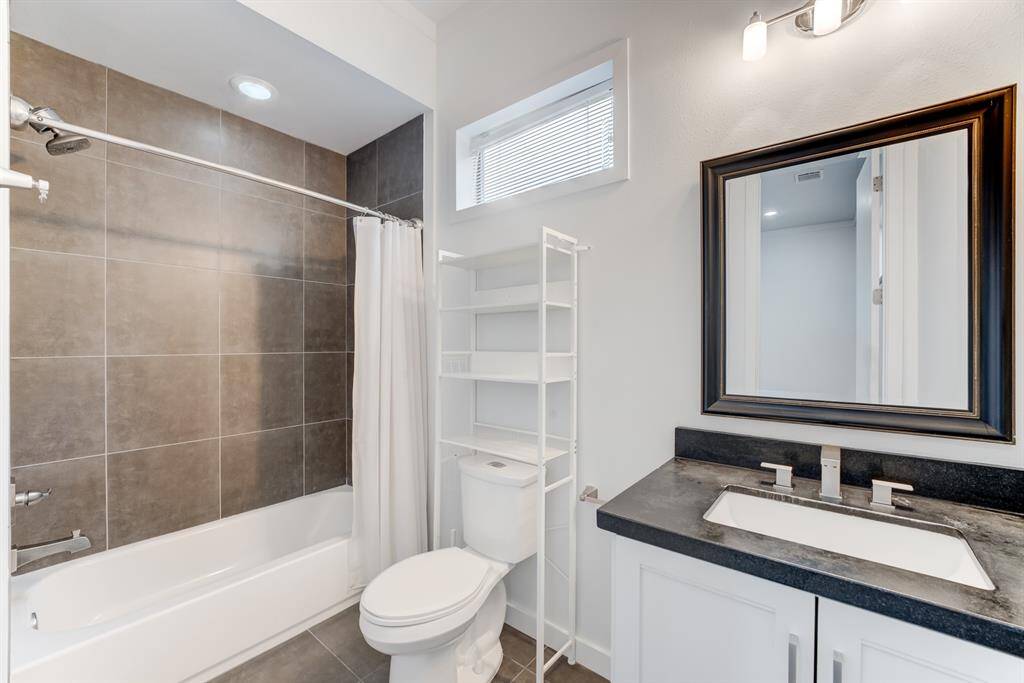 This modern bathroom features a sleek design with a large mirror, a dark countertop with a built-in sink, and a white cabinet. The shower-tub combo has a neutral tile surround, and there's a convenient open shelving unit next to the toilet for storage.
