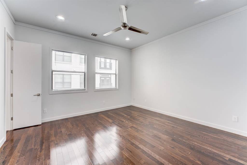 Bright, spacious room with hardwood floors, neutral walls, and ample natural light from two large windows. It features a modern ceiling fan and recessed lighting, offering a versatile space for various uses.