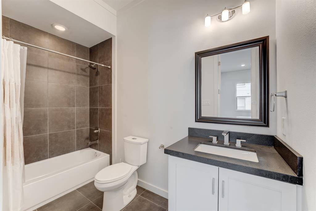 Modern bathroom with a sleek design featuring a dark countertop vanity, large mirror, and a bathtub with a tile surround. Bright and clean with contemporary fixtures.