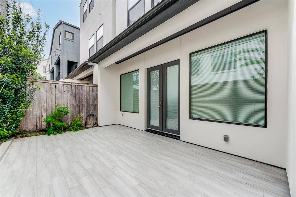 This photo showcases a modern, private patio area with sleek, light-colored tiles and large windows with blinds. The space is enclosed by a wooden fence and greenery, providing a cozy outdoor retreat.