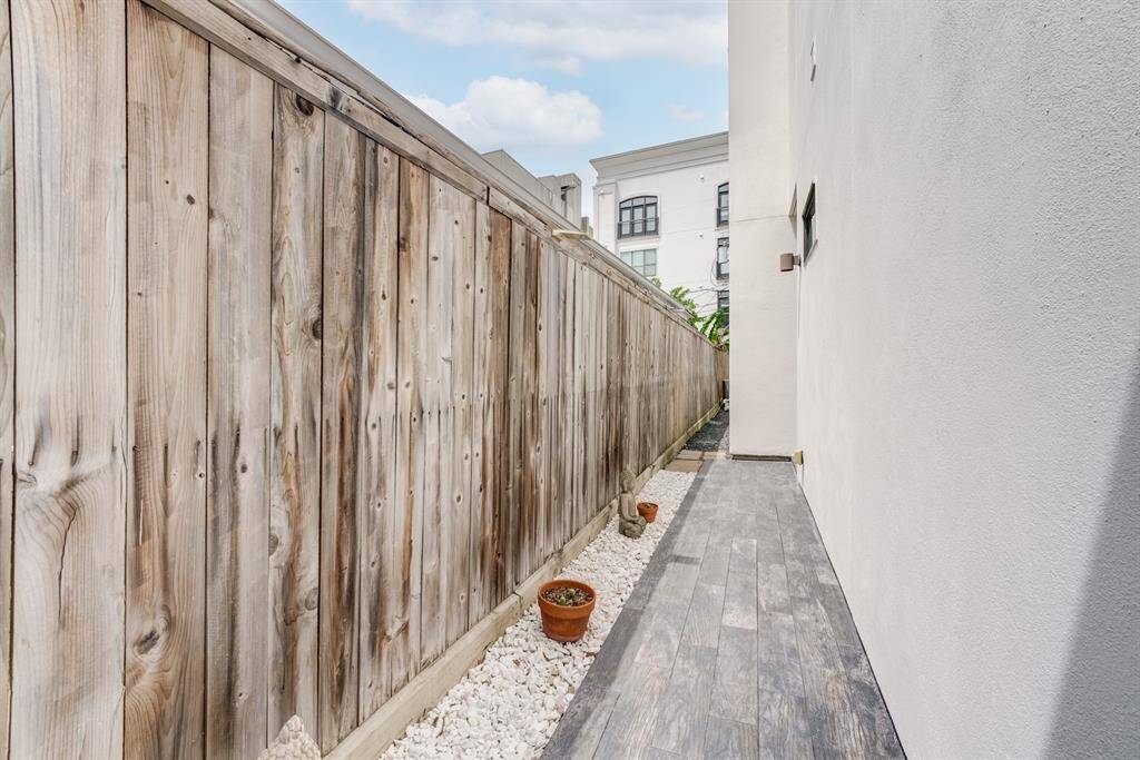 This photo shows a narrow outdoor side pathway with a wooden fence on one side and a white wall on the other. The ground features wood-like tiles and white gravel with small potted plants, offering a clean and low-maintenance area.