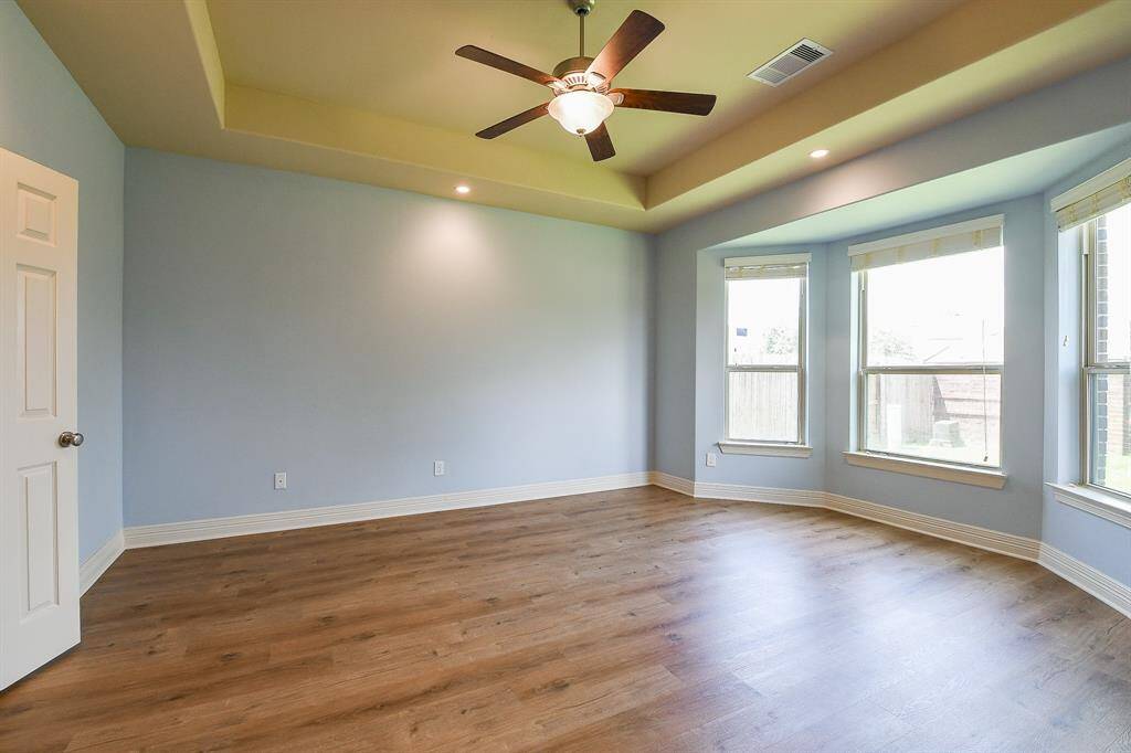 Primary bedroom features elegant LVP flooring, soft blue walls, and a tray ceiling with recessed lighting. Large windows offer plenty of natural light, complemented by a stylish ceiling fan. Ideal for a cozy, inviting atmosphere.