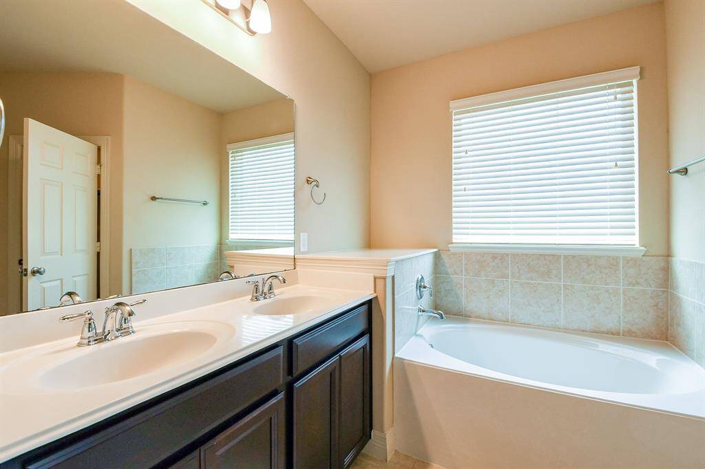 This bathroom features a dual-sink vanity with chrome fixtures, a large mirror, and a soaking tub with tiled surround. There's ample natural light from the window with blinds.