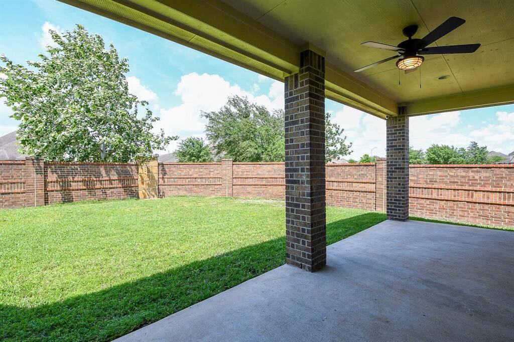 A spacious covered patio with a ceiling fan overlooks a private, fenced backyard with lush green grass, perfect for outdoor relaxation and entertainment.