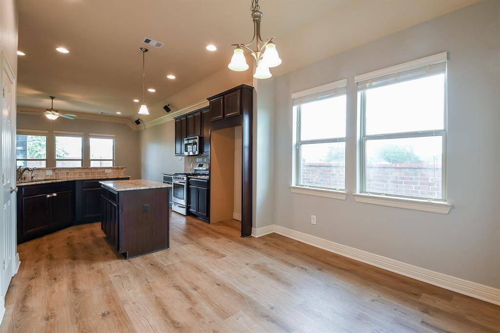 Open kitchen and dining area with new flooring. The kitchen features dark cabinetry, stainless steel appliances, and an island with a granite countertop. Large windows offer natural light, and recessed lighting adds a modern touch.
