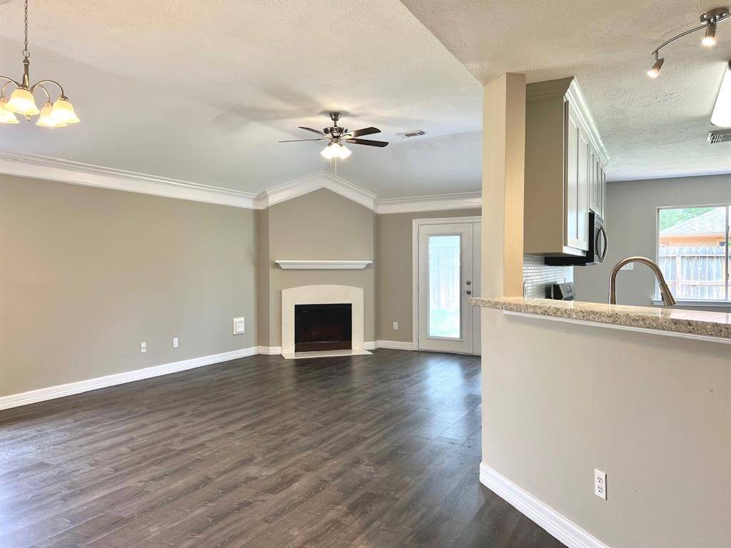 The photo showcases a spacious open-plan living area with wood flooring, a cozy corner fireplace, and a ceiling fan. The kitchen features a breakfast bar, modern cabinetry, and stainless steel appliances. A door leads to the backyard, providing natural light and outdoor access.