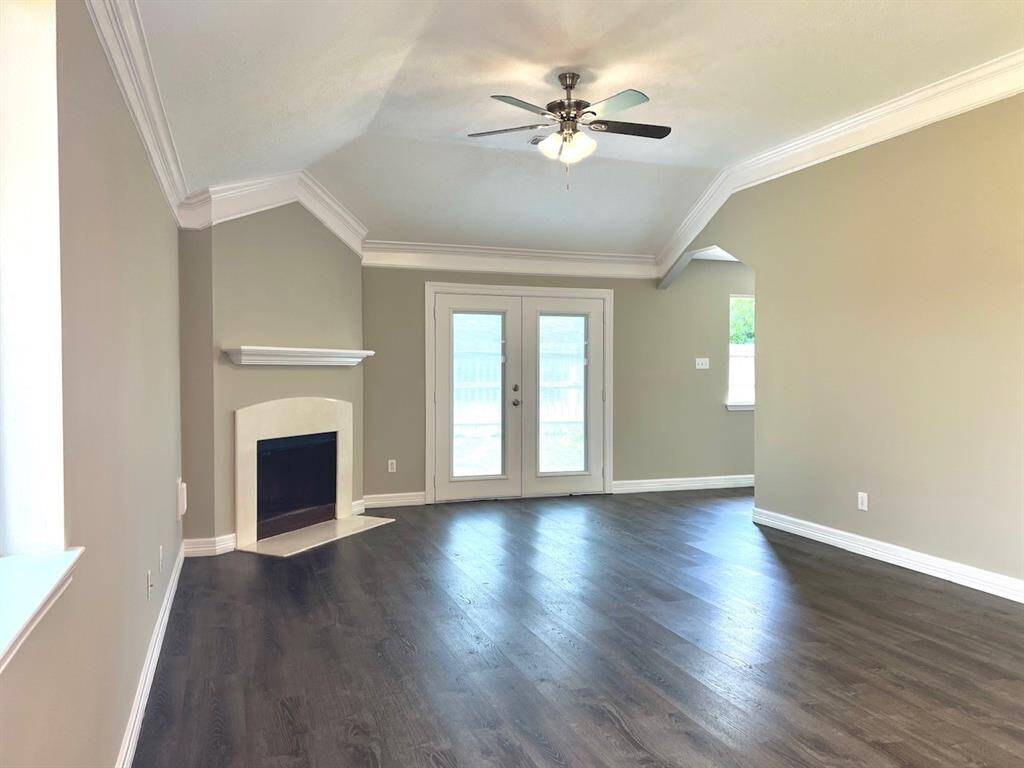 This spacious living room features elegant dark wood flooring, a cozy corner fireplace, and abundant natural light. The room includes a ceiling fan and French doors leading to the outside, offering a perfect blend of comfort and style.
