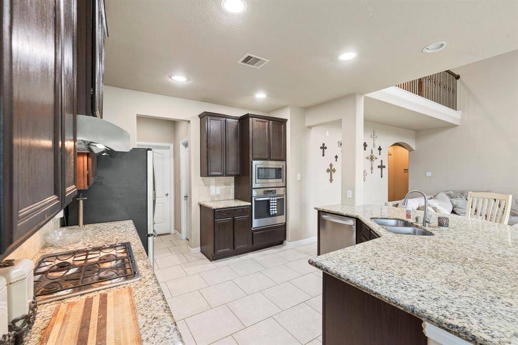 This kitchen features dark wood cabinets, granite countertops, and stainless steel appliances, including a built-in oven and microwave. The open layout leads into the living area, complemented by tile flooring and recessed lighting for a modern touch.