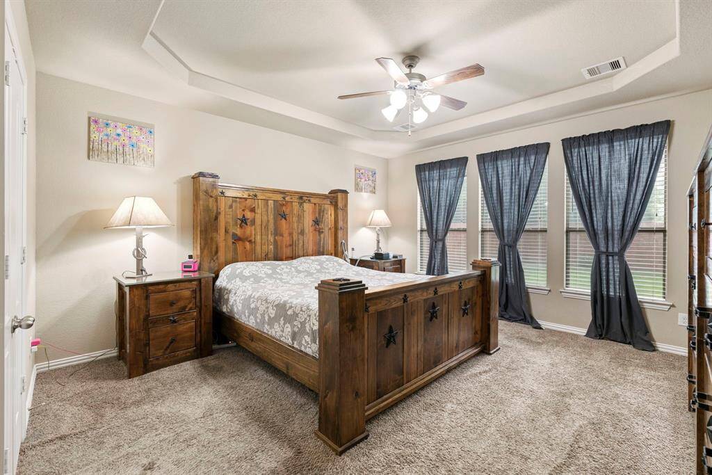 This primary bedroom features a complemented by a tray ceiling and ceiling fan. It has three large windows providing ample natural light, and cozy carpeting.