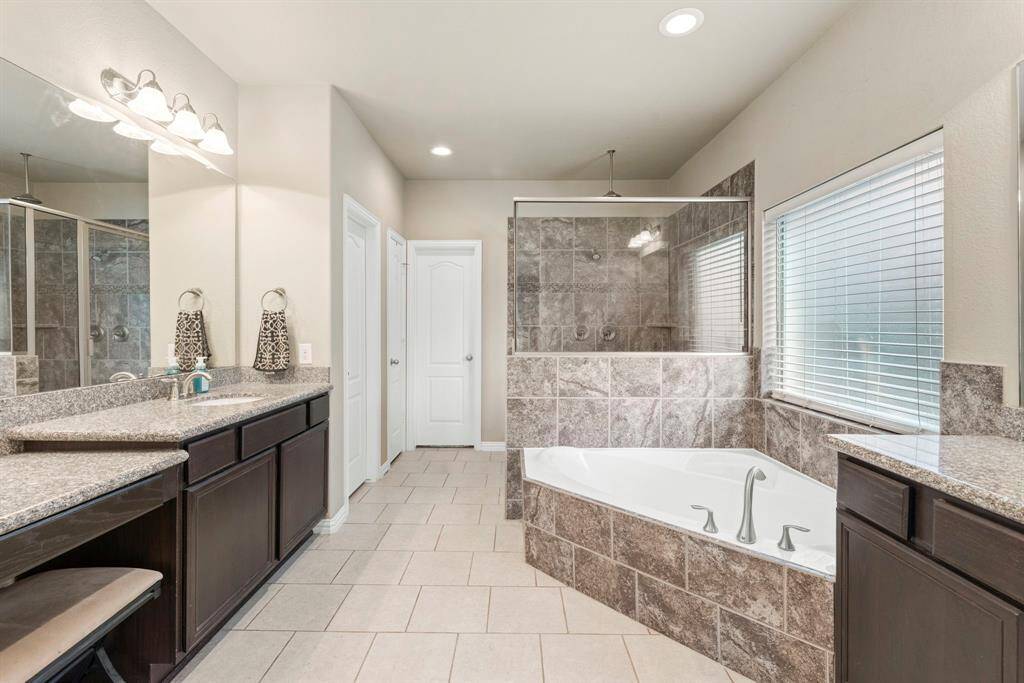 This spacious bathroom features a luxurious corner bathtub, a large tiled shower, and dual topped vanities with ample storage, all enhanced by natural light from a sizable window.