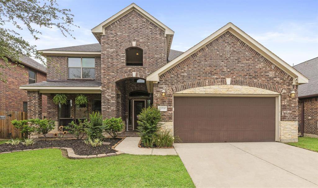 This photo showcases a charming two-story brick home with a well-maintained front yard. The house features a two-car garage, a welcoming front porch, and attractive landscaping, set against a serene blue sky.