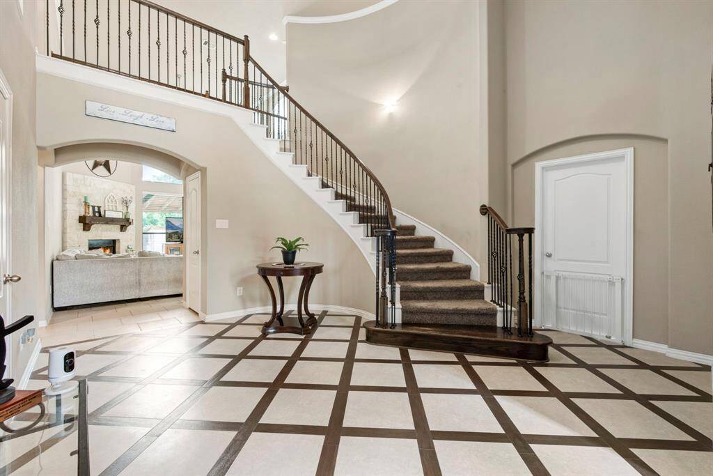 This elegant entryway features a grand staircase with wrought iron railings, a stylish tiled floor with a geometric pattern, and leads to a spacious living area with a cozy fireplace.