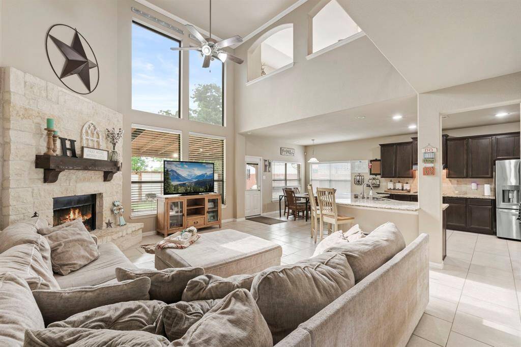 This spacious living area features high ceilings and large windows that provide natural light. It includes a cozy stone fireplace, an open layout with a seamless flow into the dining and modern kitchen areas, and a comfortable seating arrangement. Ideal for family gatherings and entertaining.