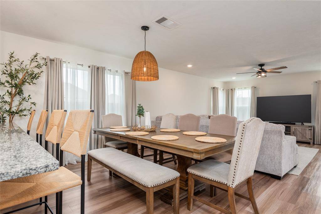 Spacious open-concept dining and living area featuring a large wooden table, upholstered chairs, and a cozy seating area with a TV. The space is bright with natural light, complemented by light wood flooring and a stylish pendant light.
