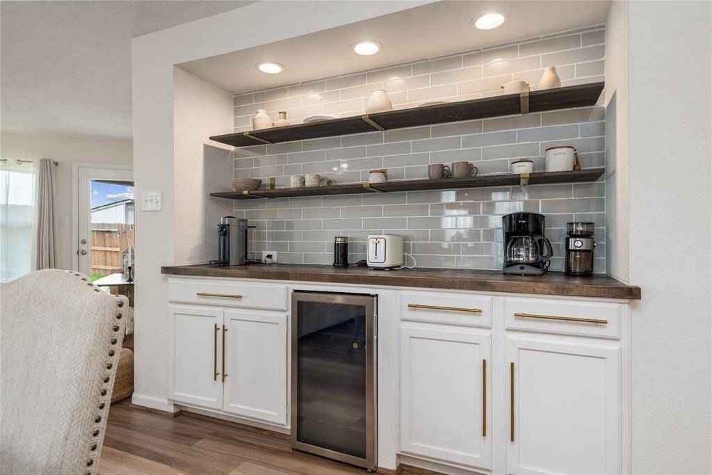 This photo showcases a modern coffee nook with sleek, open shelving, a stylish tile backsplash, and elegant white cabinetry. It features a mini fridge, countertop appliances, and ample storage, creating a functional and inviting space.