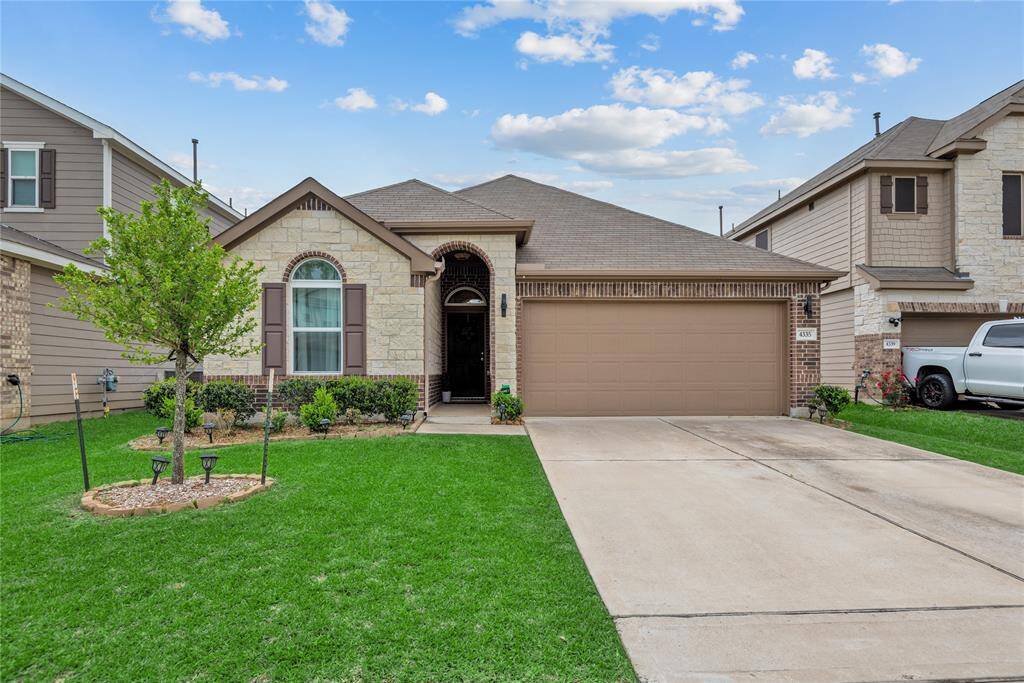 Cozy single-story home with a charming stone and brick exterior. Features an arched entry, two-car garage, and a well-maintained front yard with a young tree and landscaped garden beds.