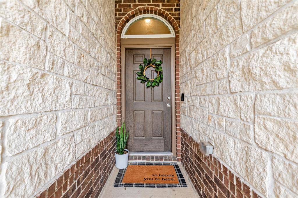 Welcoming entrance with a rustic wood door framed by stone walls. Features a charming wreath, cheerful doormat, and a potted plant, creating a warm first impression.