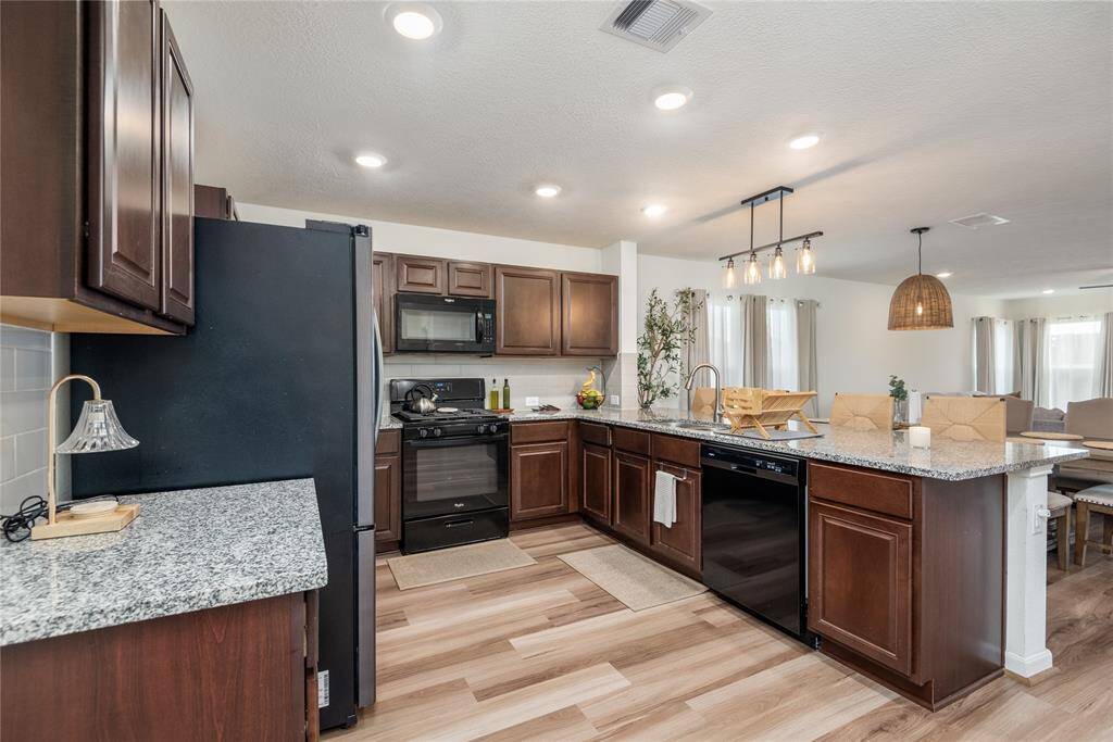 This modern kitchen features dark wood cabinets, granite countertops, and stainless steel appliances. It opens into a spacious living area with ample natural light and stylish light fixtures, perfect for entertaining.