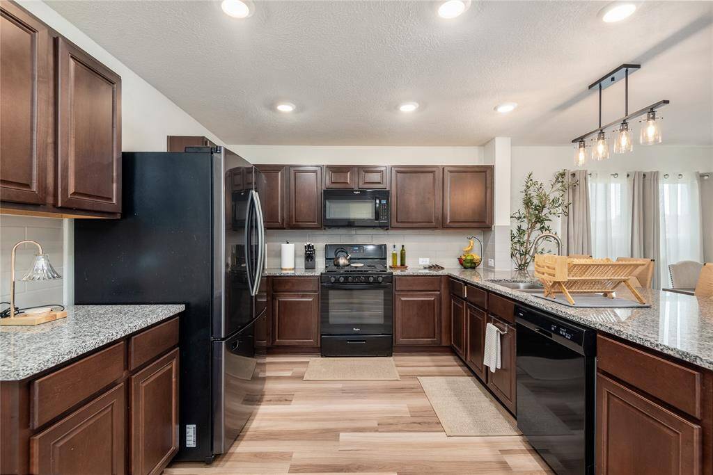 This kitchen features modern stainless steel appliances, dark wood cabinetry, and granite countertops. The open layout includes an island with a sink and stylish pendant lighting, complemented by light wood flooring.