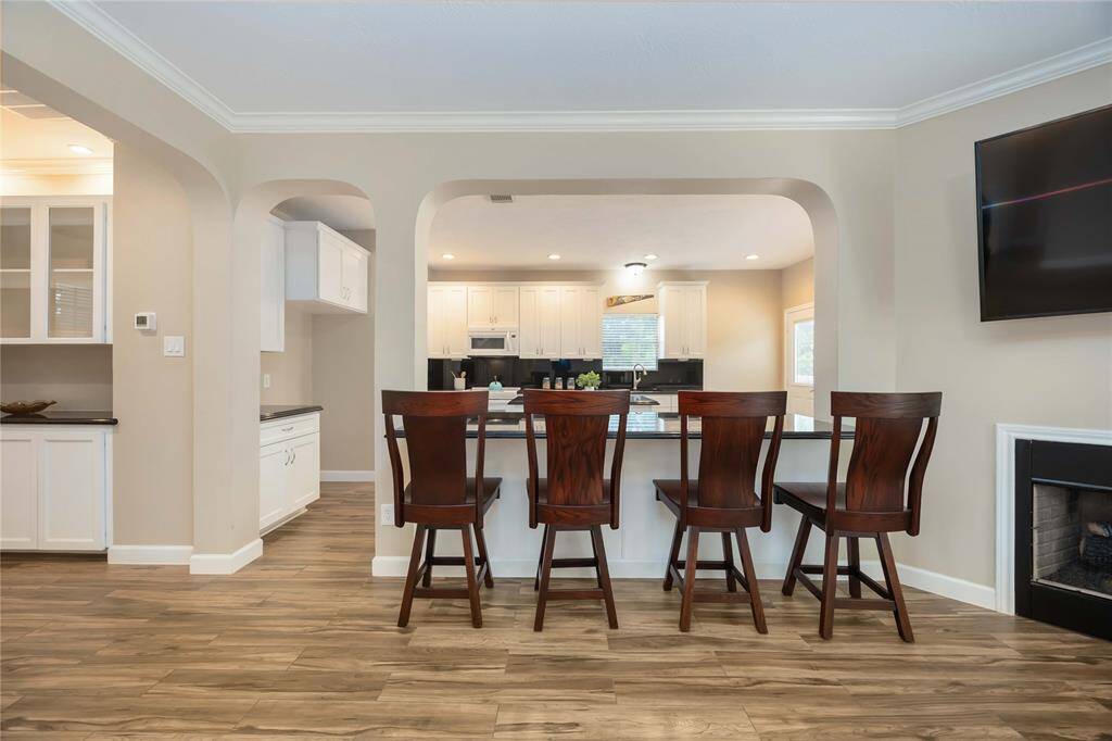 An open-concept kitchen with a breakfast bar featuring four wooden stools. The space is bright with modern white cabinetry, a sleek countertop, and a visible living area with a fireplace. The flooring is a warm wood finish, enhancing the cozy, inviting atmosphere.