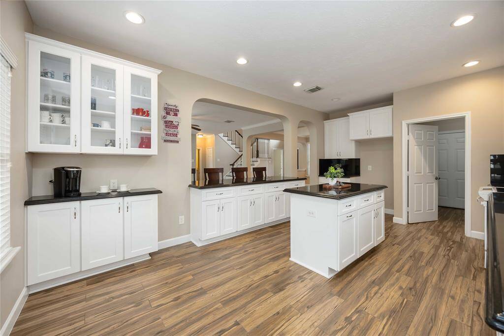 This kitchen features a modern design with hardwood floors, white cabinetry, and dark countertops. It includes a central island, recessed lighting, and open shelving, creating a bright and welcoming space. The layout offers easy access to adjacent rooms through open archways.