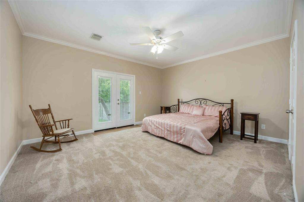 Spacious primary bedroom with neutral walls and carpet, featuring a ceiling fan for comfort. It includes a decorative bed and side tables, a rocking chair, and French doors leading to an outdoor space.