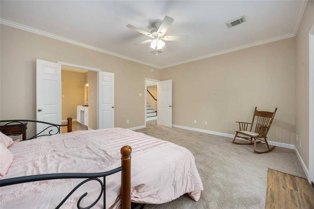This spacious primary bedroom features neutral walls, carpet flooring, and a ceiling fan. It includes an ensuite bathroom with double doors and a cozy seating area with a rocking chair.