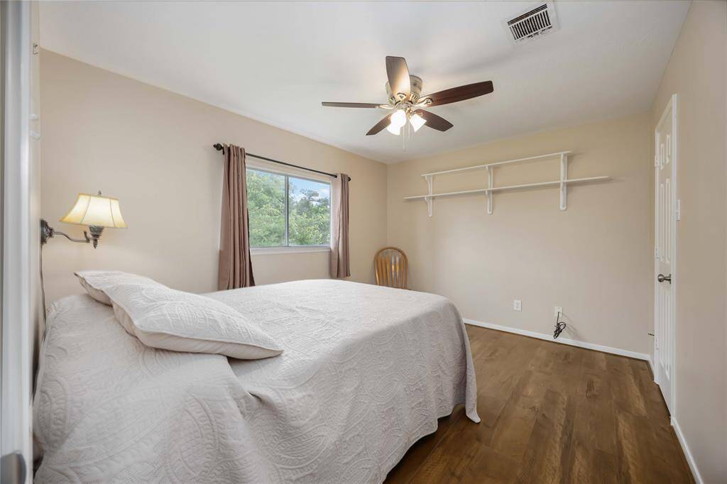 This bedroom features a cozy, neutral-toned space with a ceiling fan, wood flooring, and a large window with a view. It includes a wall-mounted lamp, shelf for storage, and ample natural light.