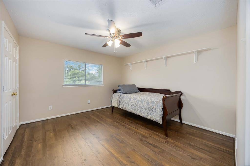 This room features a warm wood floor, neutral walls, and a ceiling fan. A window provides natural light, and a shelf offers extra storage. It's currently furnished with a daybed, making it versatile for a bedroom or office.
