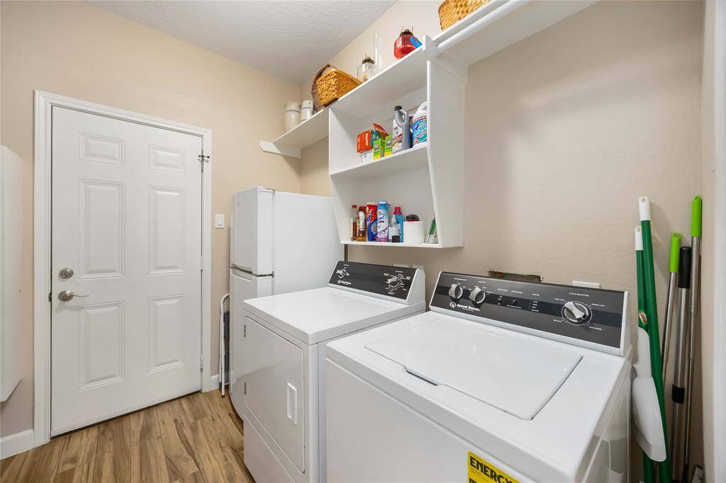 This laundry room with a washer and dryer, a white storage shelf above for cleaning supplies, and a utility closet. The room has a clean, modern look with wood-style flooring and neutral walls.