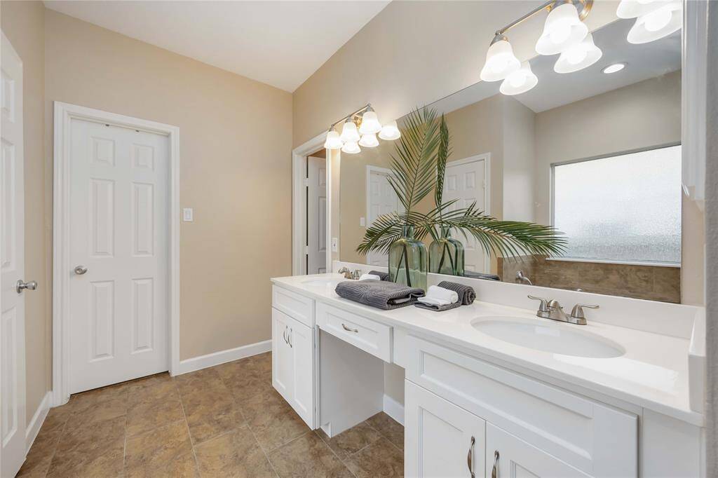 Bright and spacious bathroom with a large mirror, dual sinks, and modern lighting. It features elegant cabinetry, a frosted window for privacy, and a decorative plant for a fresh touch.