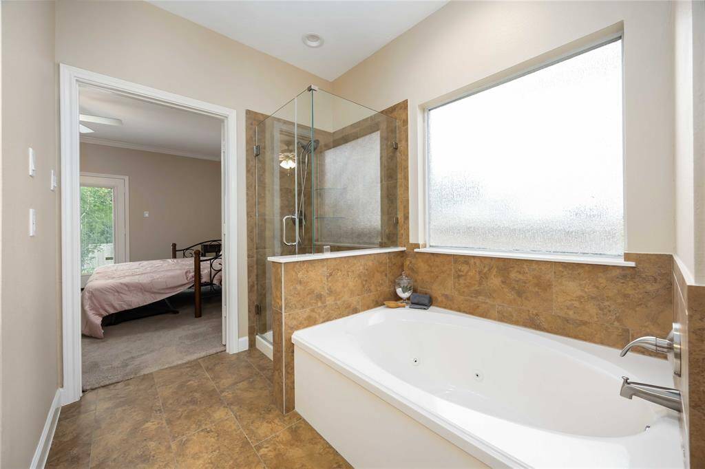 Bright, modern bathroom with a spacious bathtub, glass-enclosed shower, and a large frosted window for natural light. It opens into a cozy bedroom with a view of greenery outside.