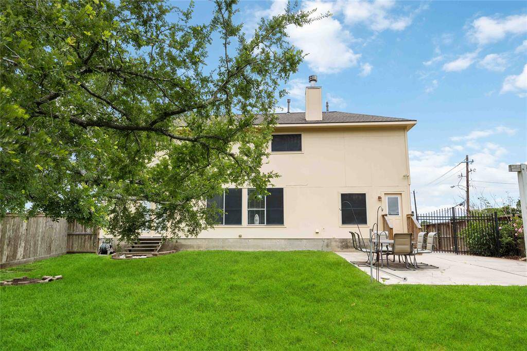 The backyard of a two-story home with a large green lawn, a spacious patio area with outdoor furniture, and a mature tree providing shade. The space is ideal for relaxation and entertaining.