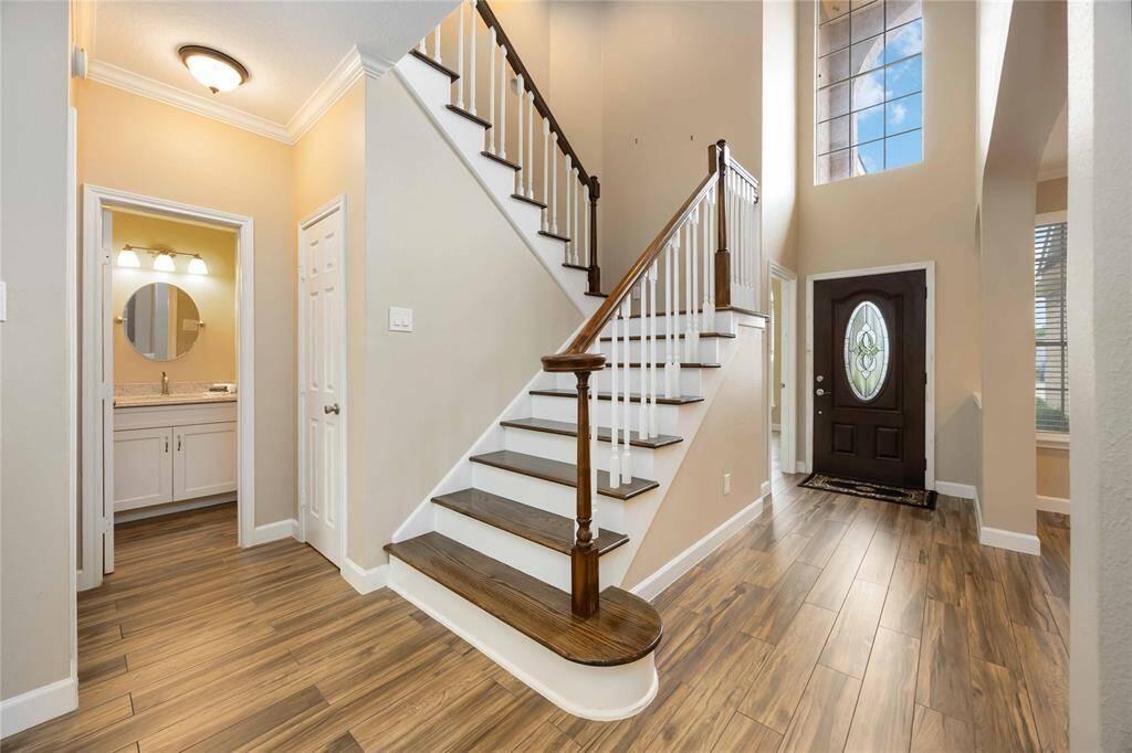 This photo showcases a bright, welcoming entryway featuring a wooden staircase with white railings, high ceilings, and a large window. The space includes wood-look flooring and leads to a bathroom with a modern vanity.