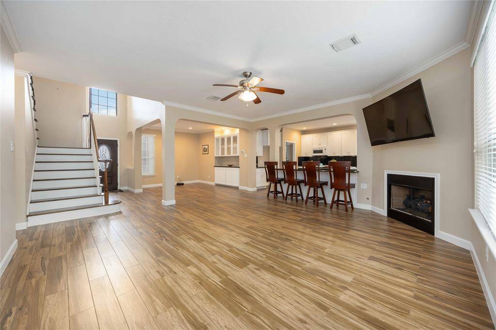 A spacious, open-concept living area with warm wood flooring, a modern kitchen with bar seating, a cozy fireplace, and a wall-mounted TV. Large windows allow plenty of natural light, and a staircase leads to the upper floor.